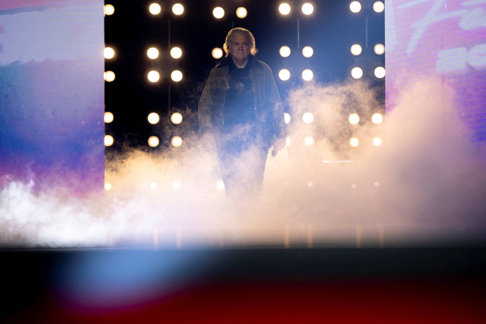 Um artista caminha em direção ao público, cercado por luzes brilhantes e uma névoa no chão. O fundo é composto por painéis iluminados que criam um ambiente dramático