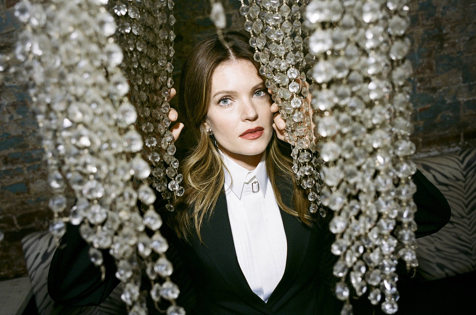 A imagem mostra uma mulher com cabelo ondulado, usando um blazer preto e uma camisa branca. Ela está posicionada entre cortinas feitas de cristais brilhantes, que criam um efeito de luz ao seu redor. O fundo é de uma parede de tijolos expostos, e o ambiente parece ser um espaço interno elegante.