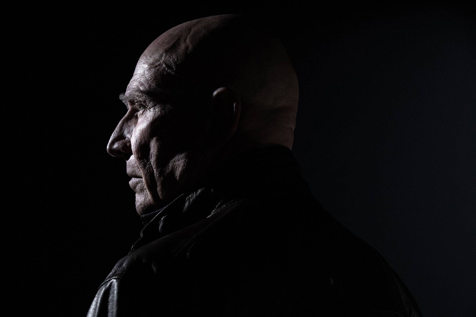 A imagem mostra a silhueta de um homem de cabeça calva, visto de perfil. Ele está usando uma jaqueta escura e a iluminação é dramática, destacando os contornos de seu rosto e a textura da pele. O fundo é escuro, criando um contraste forte com a figura do homem.