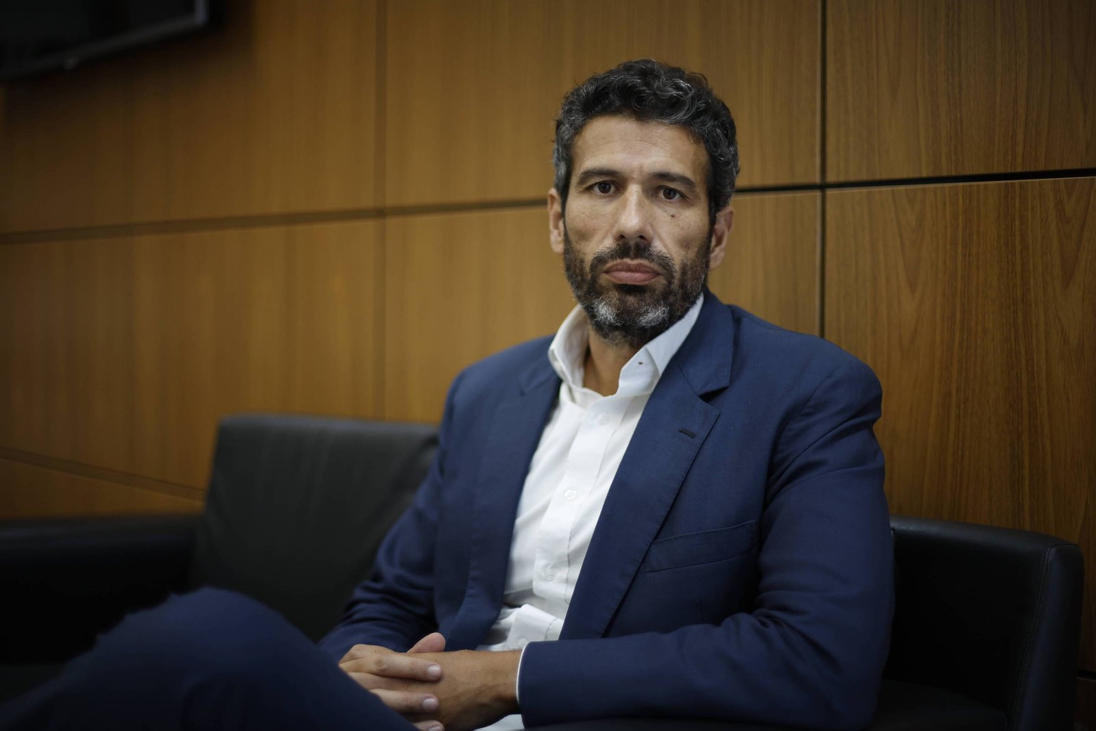 Um homem com cabelo escuro e barba, vestido com um terno azul e uma camisa branca, está sentado em um sofá preto. O fundo é composto por paredes de madeira e uma tela visível ao fundo. O homem parece estar em um ambiente de escritório, com uma expressão séria.