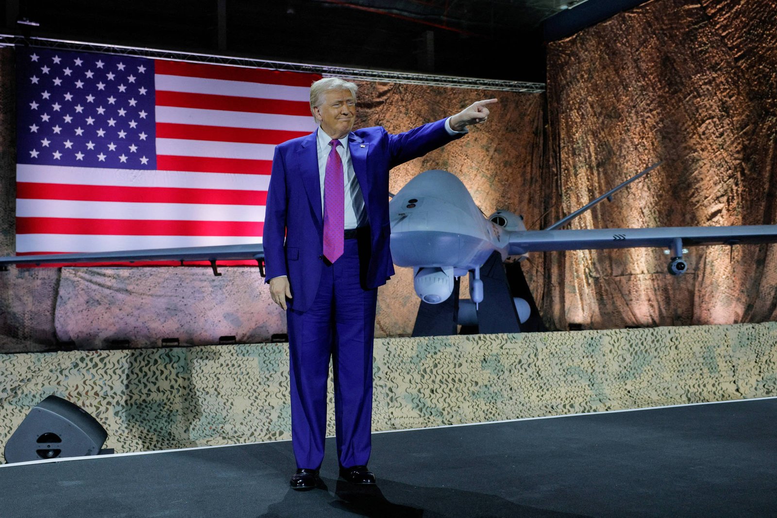 Um homem em um terno roxo está em pé em um palco, apontando para a frente. Ao fundo, há uma grande bandeira dos Estados Unidos e um drone militar em exibição. O ambiente é iluminado com luzes suaves e há uma textura de rocha na parede ao fundo.