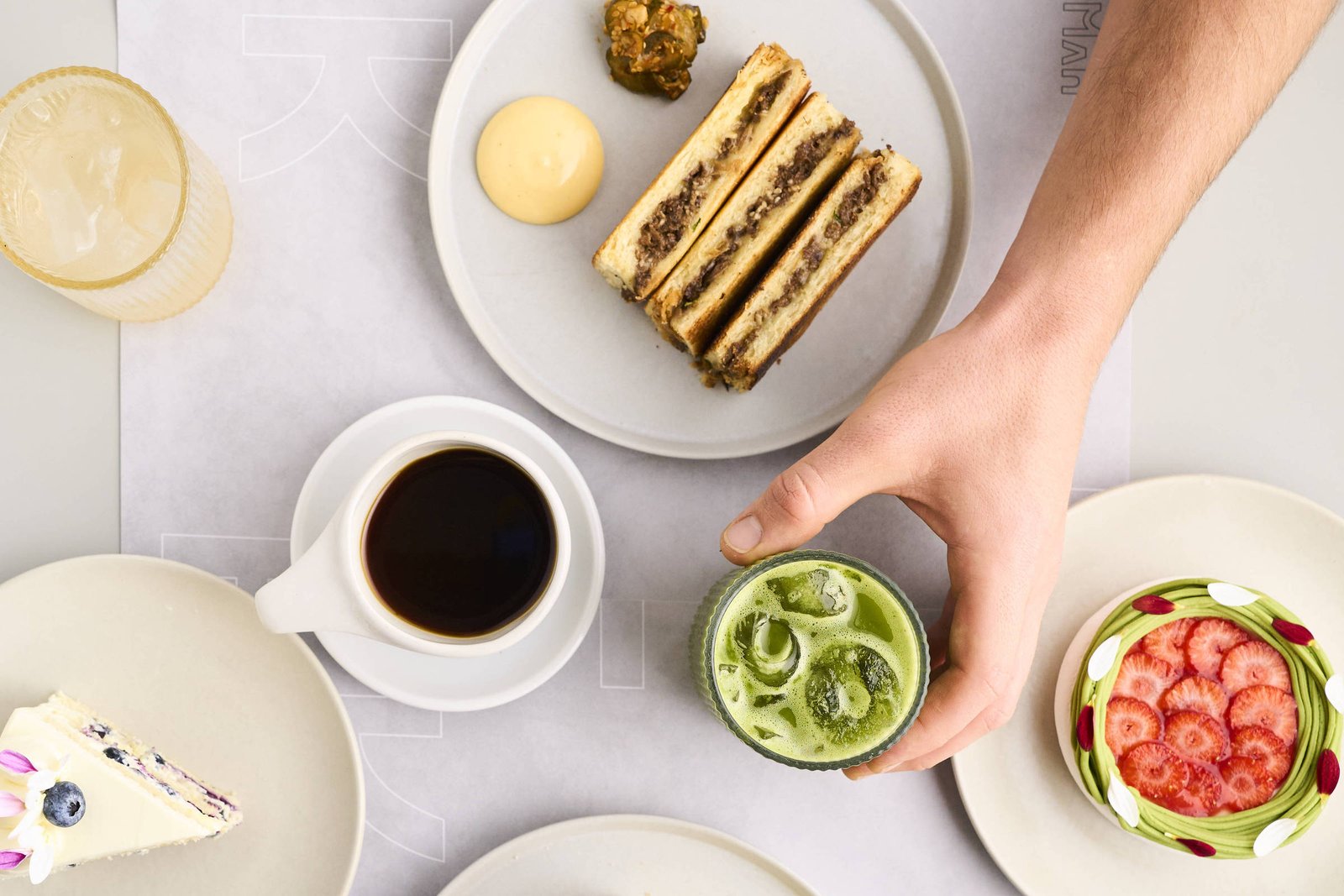 A imagem mostra uma mesa com vários pratos e bebidas. Há um prato com sanduíches, um copo com uma bebida verde, uma xícara de café, um prato com um doce e um prato com uma sobremesa colorida. As mãos de duas pessoas estão visíveis, uma segurando um sanduíche e a outra segurando o copo. O fundo é claro e a mesa é organizada.