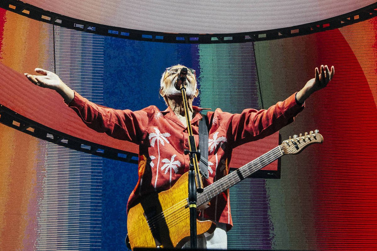 Um artista está se apresentando em um palco iluminado, com um fundo de telas que exibem padrões coloridos em espiral. Ele está usando uma jaqueta vermelha e segurando uma guitarra, com os braços abertos em uma pose expressiva. As luzes do palco criam um ambiente vibrante e dinâmico.