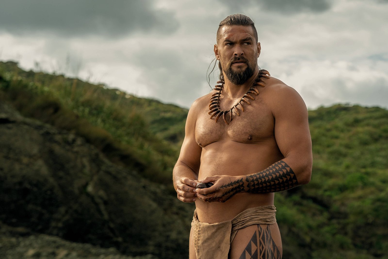 Um homem musculoso está em pé em um ambiente natural, com colinas verdes ao fundo. Ele está sem camisa, usando um colar de conchas e uma saia feita de tecido. Seu braço esquerdo possui uma tatuagem tribal. O céu está nublado, sugerindo um clima sombrio.
