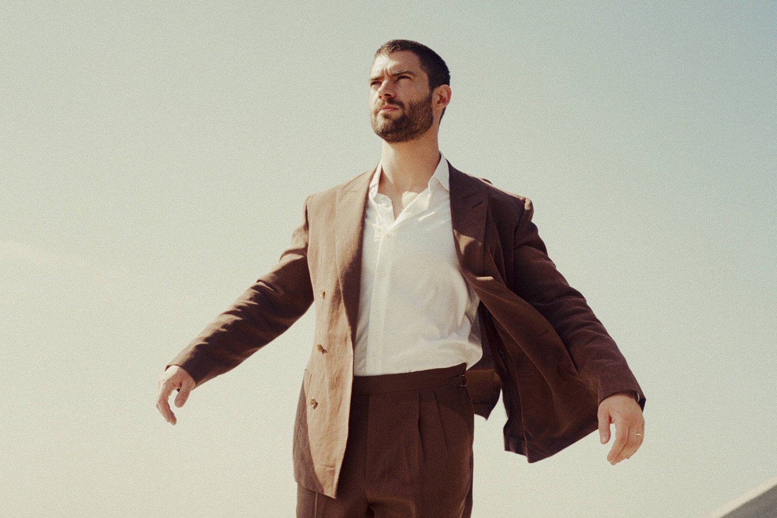 Um homem está posando em um ambiente externo, vestindo um terno marrom e uma camisa branca. Ele está caminhando com um leve sorriso, enquanto uma das mangas do terno está levemente levantada. O fundo apresenta uma vista panorâmica da cidade, com um céu claro e algumas estruturas arquitetônicas ao fundo.