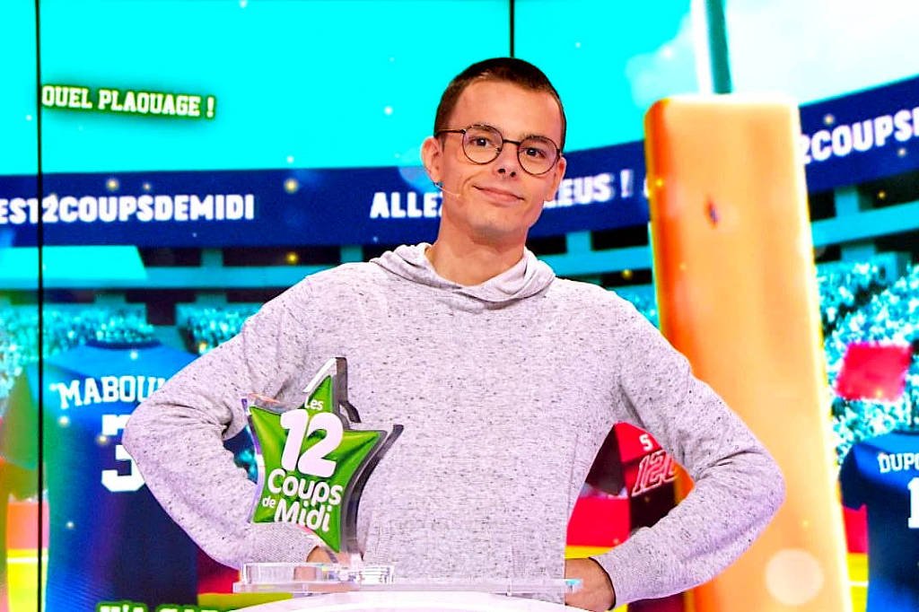 Um apresentador de programa de televisão está em pé ao lado de um troféu em forma de estrela. Ele usa um suéter cinza claro e óculos. O fundo exibe uma tela colorida com elementos relacionados a um jogo, incluindo números e palavras em francês. O ambiente parece ser um estúdio de gravação de um programa de quiz.