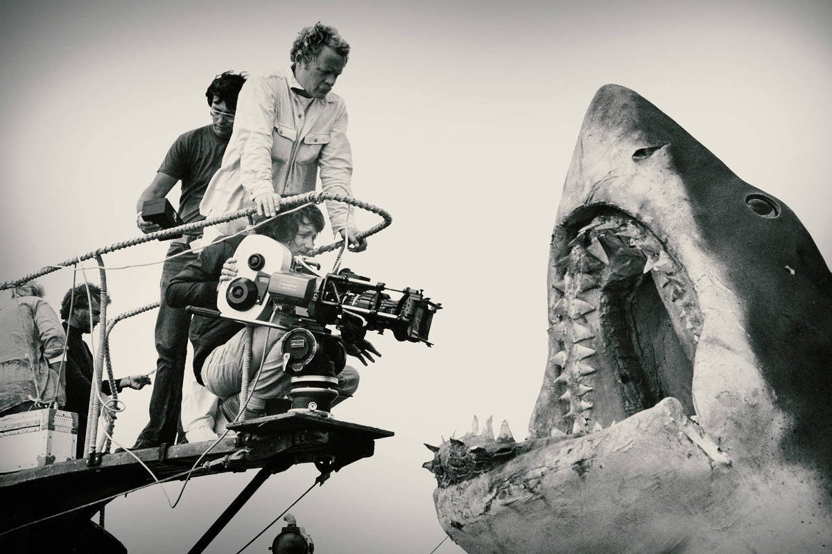 A imagem em preto e branco mostra uma equipe de filmagem em um barco, com um cinegrafista operando uma câmera voltada para um grande tubarão com a boca aberta. Há três pessoas visíveis, uma delas está segurando a câmera, enquanto as outras observam. O cenário parece ser um ambiente aquático, possivelmente uma filmagem de um filme sobre tubarões.