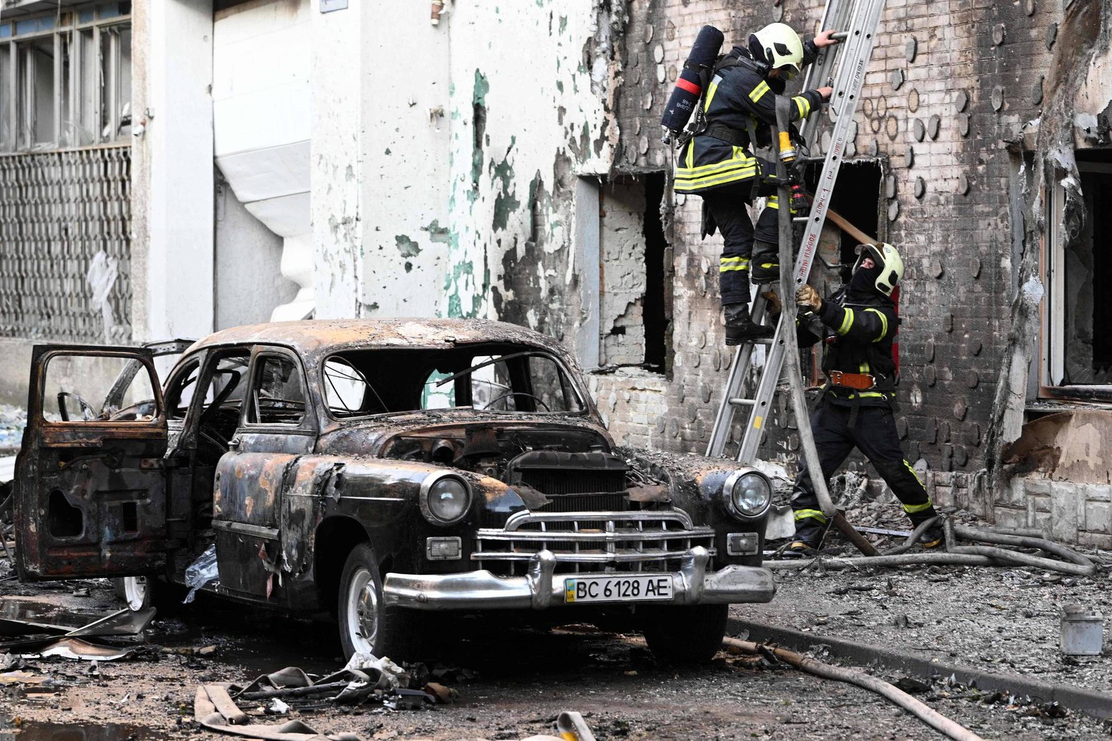 A imagem mostra um carro antigo, parcialmente queimado, estacionado em frente a um edifício danificado. O carro tem as portas abertas e está coberto de fuligem. Dois bombeiros, equipados com uniformes e capacetes, estão subindo uma escada em direção a uma janela do edifício, que apresenta marcas de incêndio e destruição. O chão está coberto de detritos e há mangueiras de incêndio visíveis ao redor.