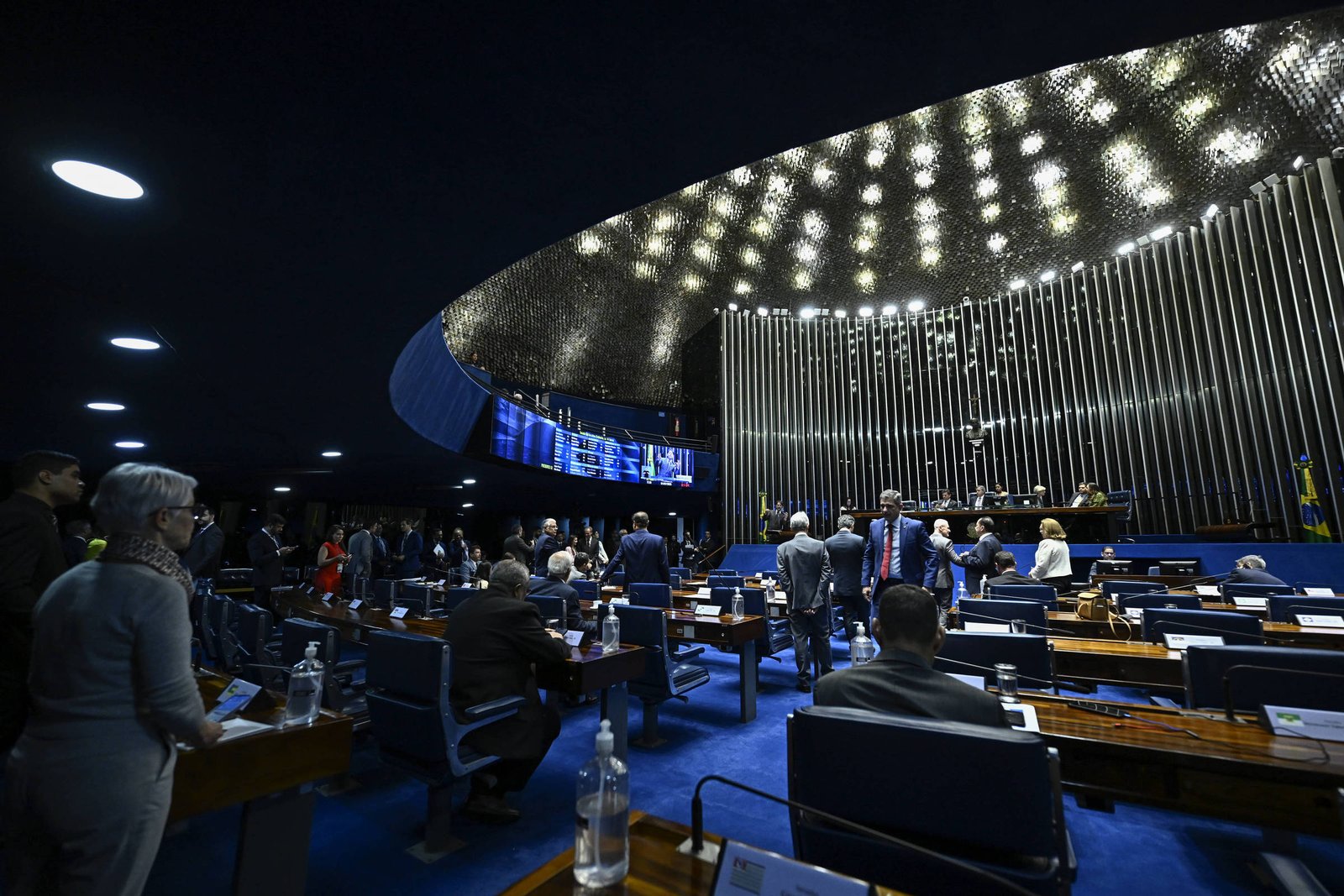 A imagem mostra o plenário do Senado Federal do Brasil, com senadores em pé e outros sentados em suas cadeiras. O ambiente é iluminado por uma iluminação moderna e o teto é curvo, com um design contemporâneo. Ao fundo, há painéis eletrônicos e a bandeira do Brasil. As mesas são de madeira e há garrafas de água sobre elas.