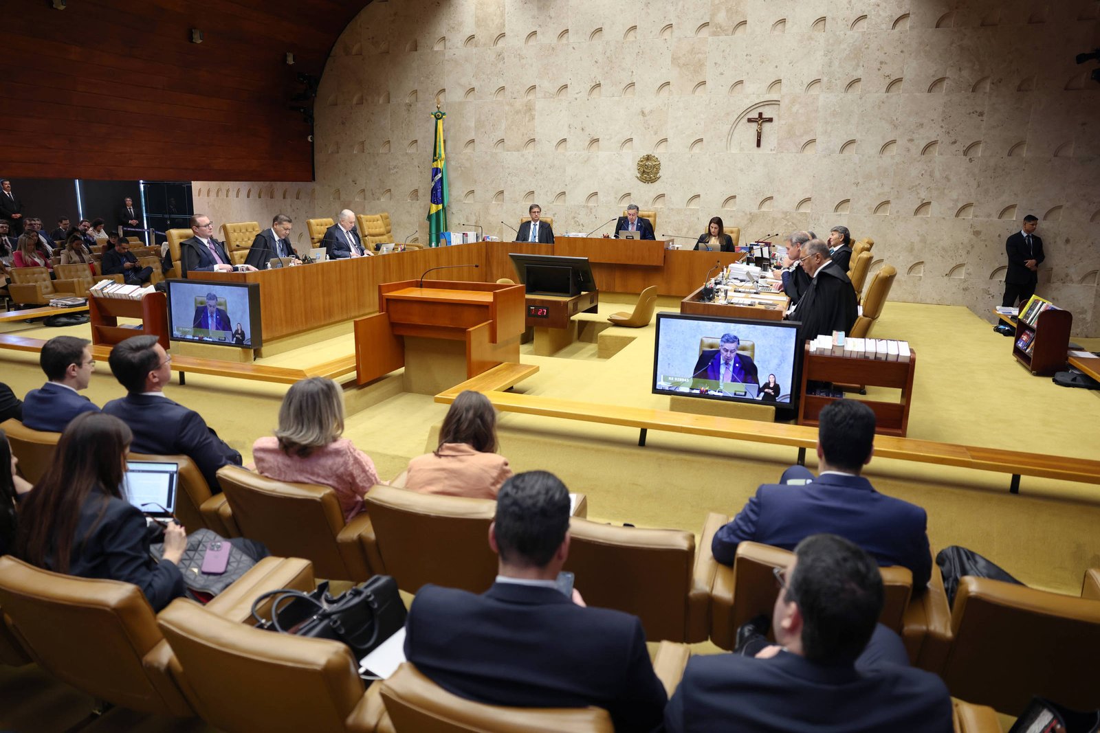 A imagem mostra uma sessão do Supremo Tribunal Federal do Brasil. No centro, há uma mesa com vários ministros sentados, alguns olhando para um telão. À frente, há um advogado ou orador falando, com um microfone à sua frente. O ambiente é bem iluminado, com paredes de tom claro e uma decoração simples. Há bandeiras do Brasil e um crucifixo na parede ao fundo. Na parte inferior da imagem, é possível ver pessoas na plateia, algumas usando dispositivos eletrônicos.