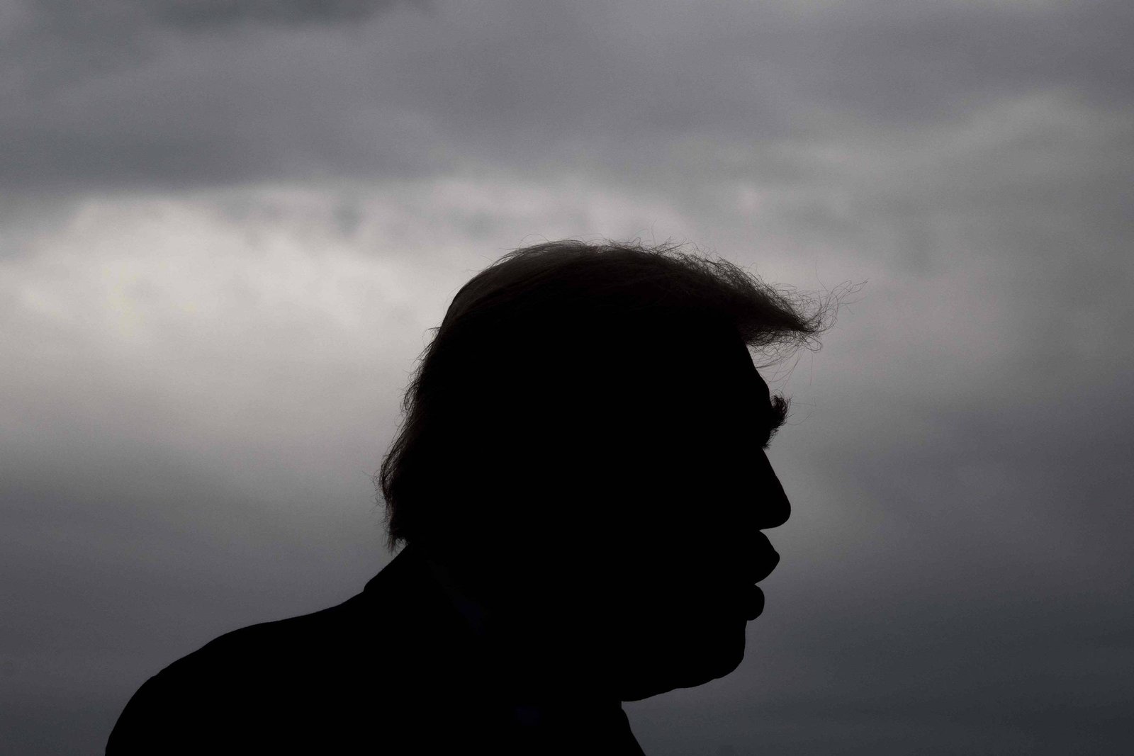 A imagem mostra a silhueta de uma pessoa contra um fundo de nuvens escuras e ameaçadoras. A figura tem um cabelo característico e está em perfil, com a boca ligeiramente aberta, sugerindo que está falando ou expressando algo. O ambiente parece sombrio devido à iluminação e às nuvens.