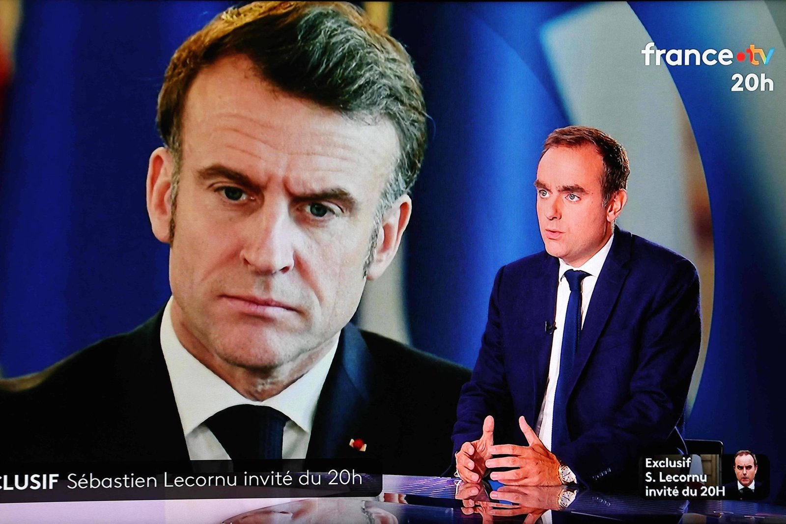 Sébastien Lecornu, vestido com terno escuro e gravata, participa de entrevista no estúdio do jornal da France 2 às 20h. Ao fundo, imagem ampliada de seu rosto aparece em tela azul.