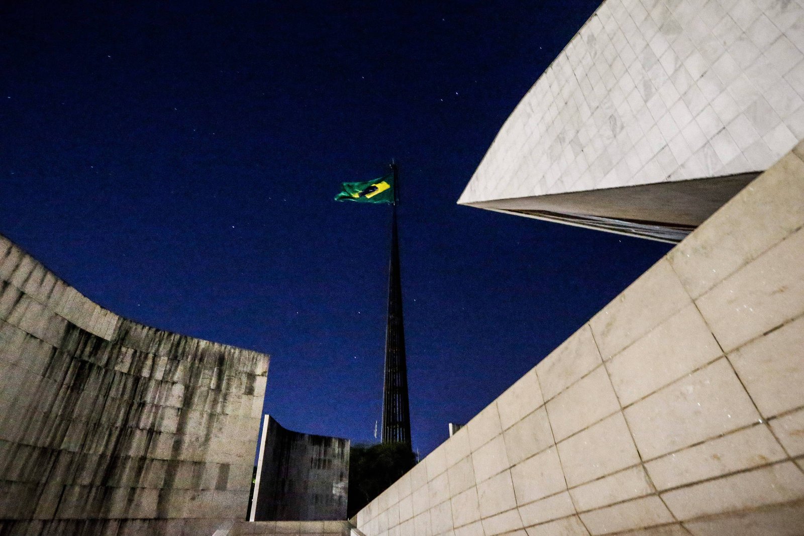 Bandeira do Brasil tremula no alto de mastro alto contra céu noturno estrelado. Estruturas arquitetônicas de concreto do Museu do Amanhã cercam a cena, com linhas angulares e superfícies claras.