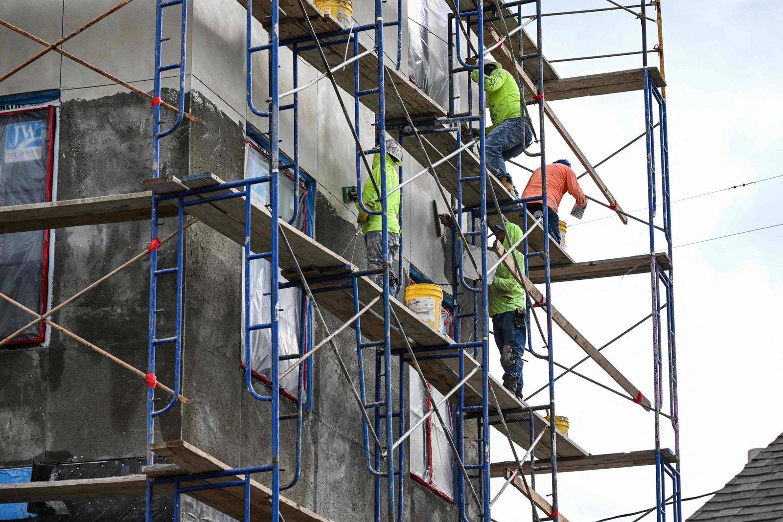 Quatro trabalhadores com coletes de segurança realizam reparos na fachada de um prédio cinza, usando andaimes metálicos azuis e pranchas de madeira. Um deles está no nível mais alto, enquanto os outros estão em níveis intermediários, com baldes amarelos próximos.