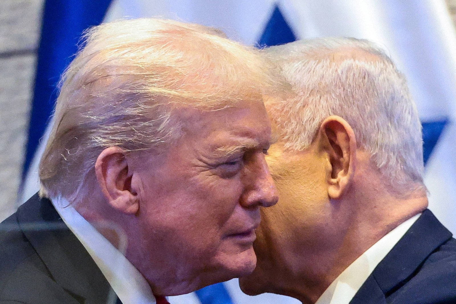 Donald Trump e Benjamin Netanyahu estão lado a lado, com Netanyahu sussurrando no ouvido de Trump. Ambos usam ternos escuros e estão diante de uma bandeira desfocada ao fundo.