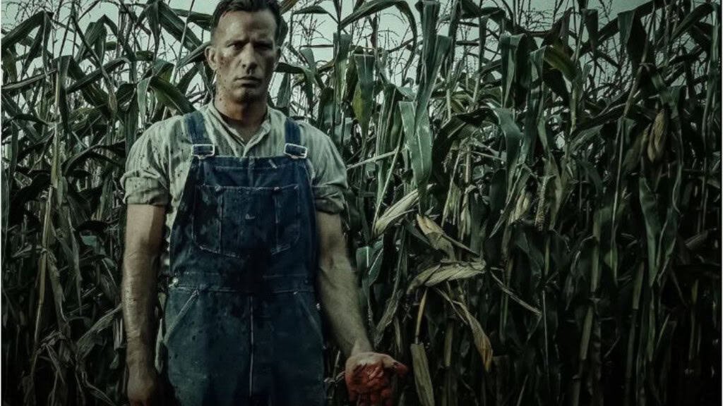 Homem com macacão e camisa, saindo do milharal com as mãos cheias de sangue.