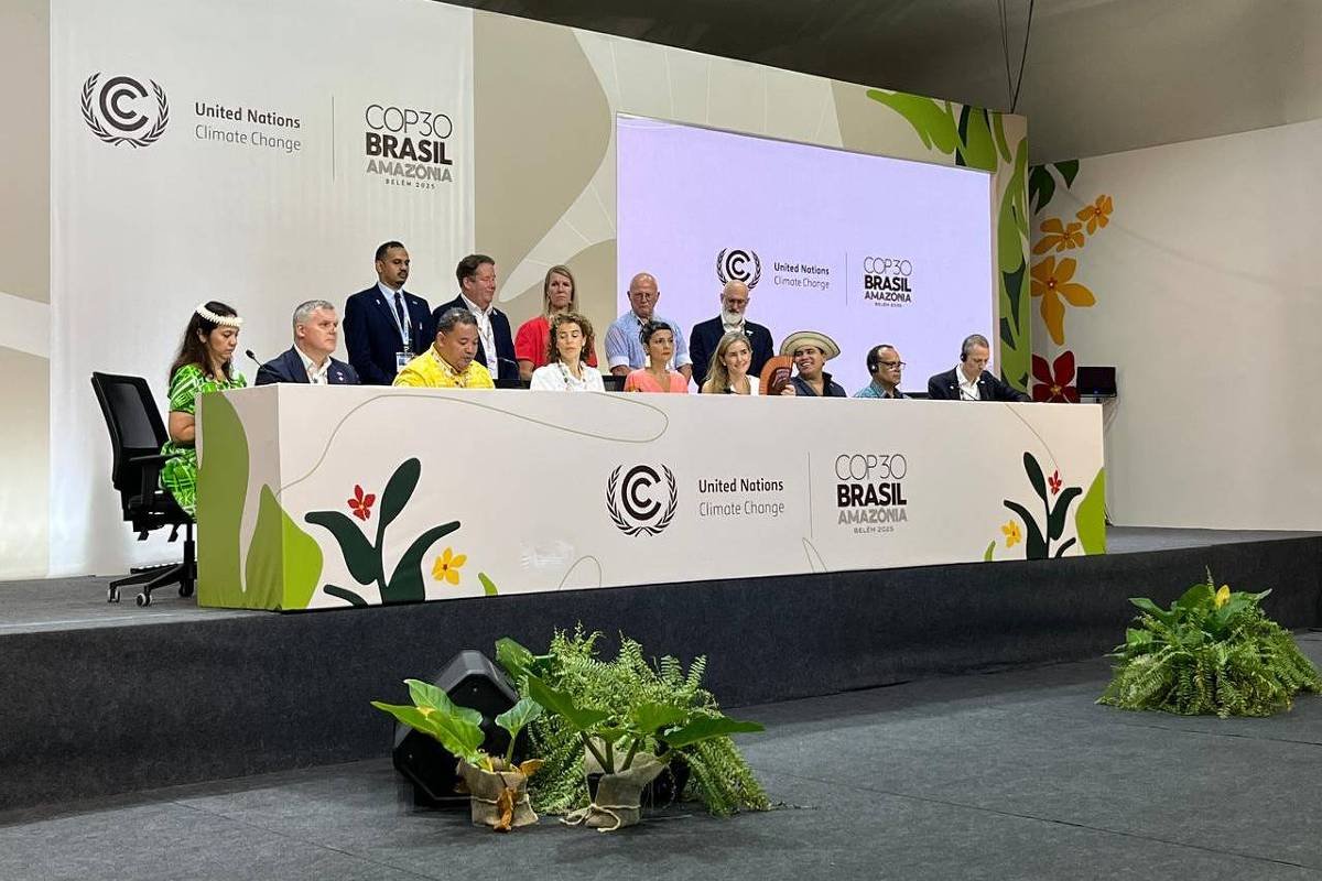 Doze delegados sentados em mesa longa com logotipos da COP30 Brasil Amazônia e ONU Mudança do Clima ao fundo. Palco decorado com plantas e iluminação de evento formal.