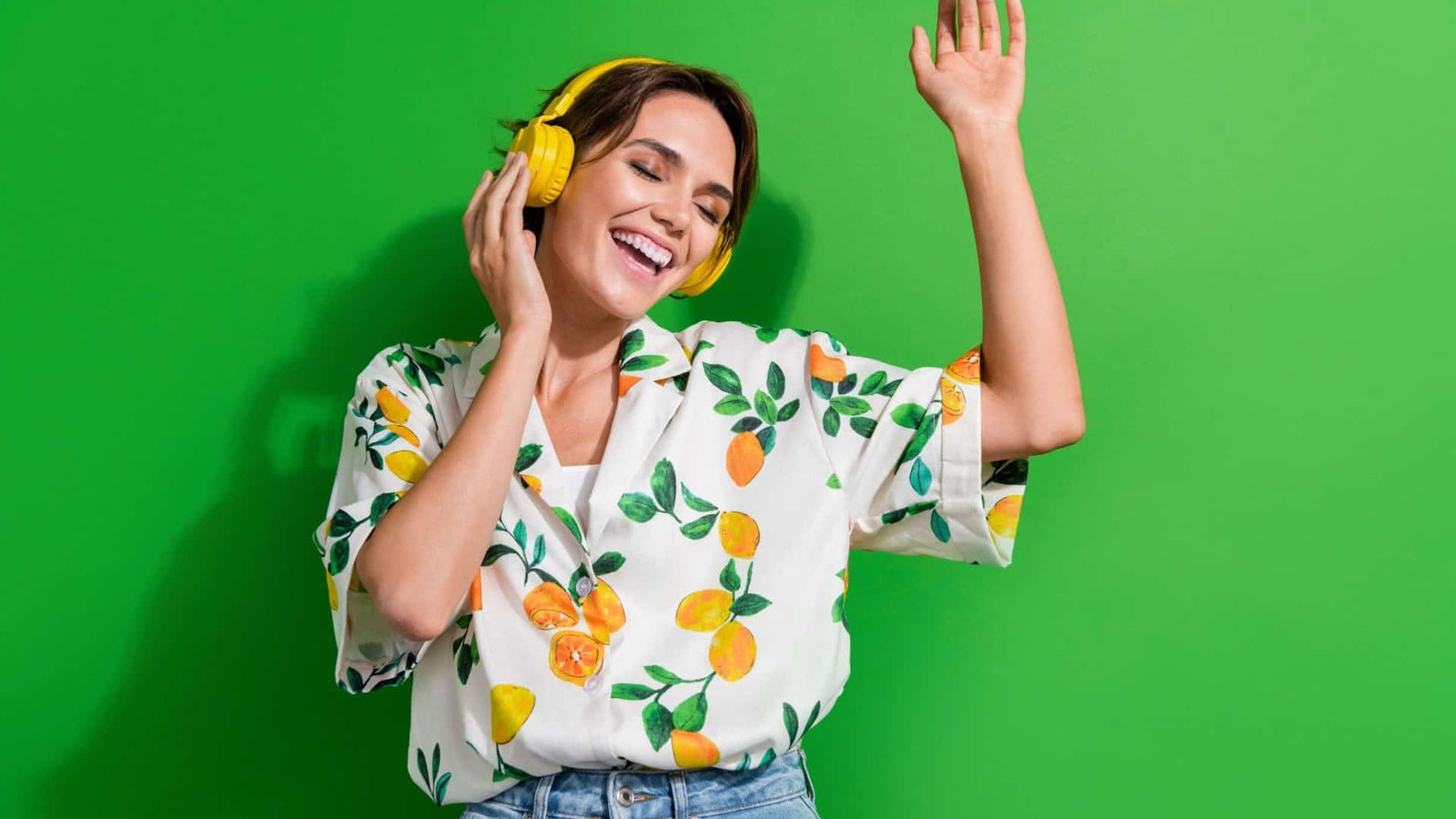 Mulher de cabelo curto e castanho, sorrindo com os olhos fechados, curte música com fones amarelos vibrantes. Veste camisa branca estampada com limões e folhas, calça jeans, e está diante de um fundo verde intenso. A pose transmite alegria e leveza.