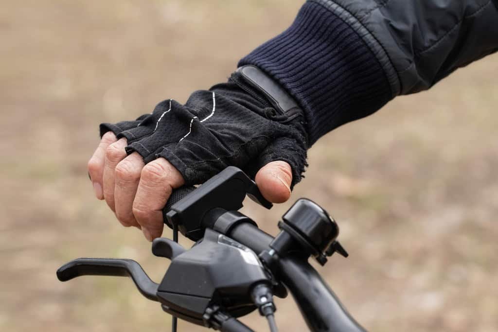 Close-up da mão de um ciclista usando luvas pretas pressionando o acelerador de polegar no guidão de uma bicicleta elétrica. O foco está no mecanismo de aceleração manual.