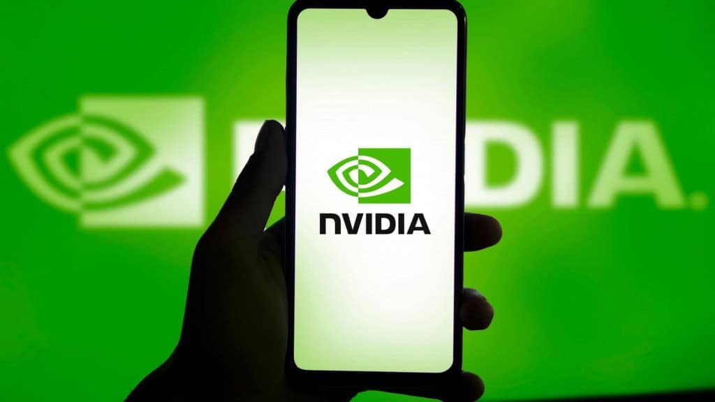 Logo da Nvidia ao fundo; à frente, uma mão segurando um celular com o logo da empresa em sua tela