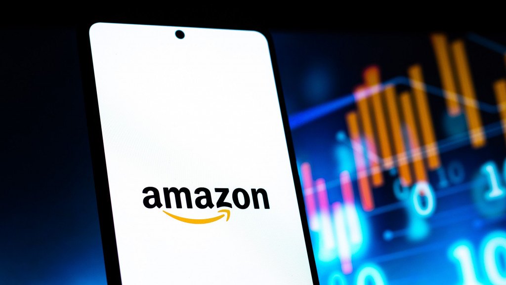 Atrás, desfocados, gráficos financeiros; à frente, logo da Amazon na tela de um smartphone