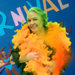Myrinha Vasconcellos sorrindo com plumas amarelas em frente a painel azul decorado com letras coloridas formando a palavra “Carnaval”.