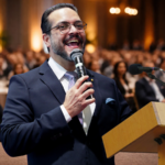 Mestre de cerimônias conduzindo evento corporativo com microfone em um auditório cheio, público ao fundo desfocado e ambiente elegante.