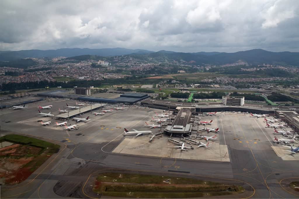 Vista aérea de aeroporto com aviões estacionados no pátio e terminal ao fundo sob céu nublado