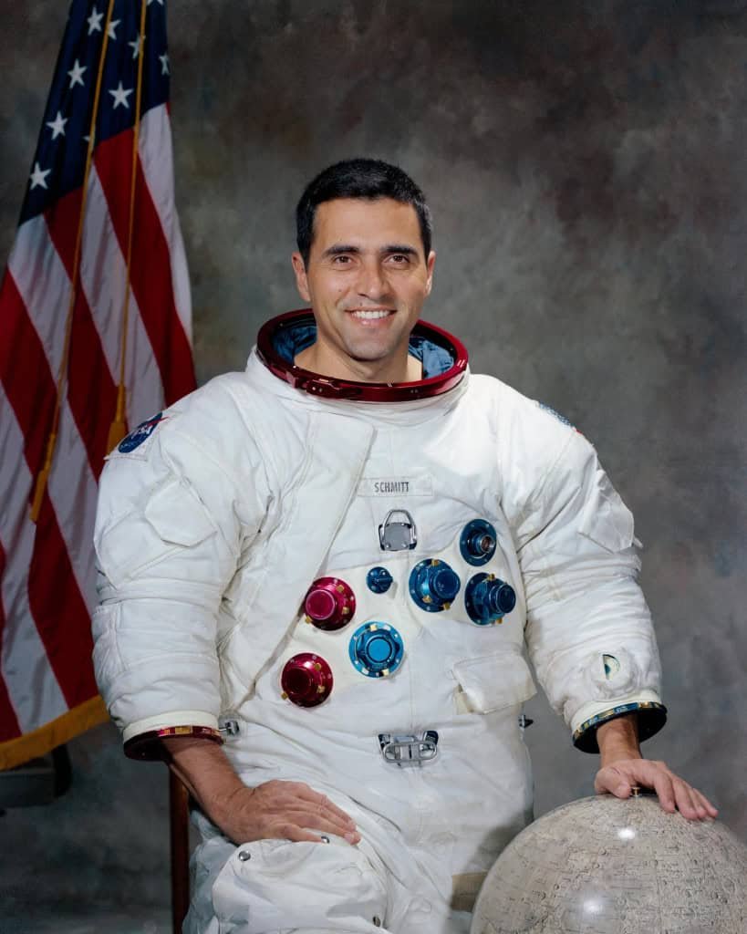 Harrison Schmitt em traje espacial branco posa em estúdio ao lado de uma bandeira dos Estados Unidos e um globo lunar