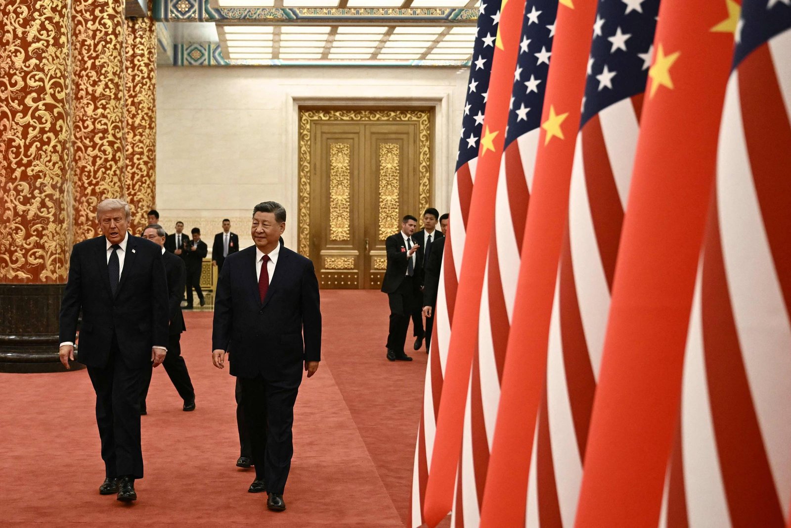 Dois líderes, um dos Estados Unidos e outro da China, caminham lado a lado em um salão com tapete vermelho. À direita, fileira de bandeiras dos EUA e da China alternadas. Ao fundo, portas douradas e seguranças observam a cena.